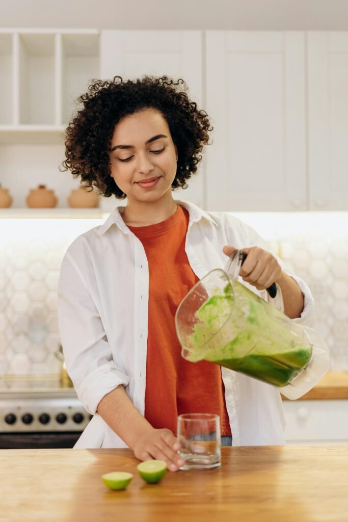 pexels-photo-6707376-6707376 Woman in casual attire pouring a fresh green smoothie into a glass in a modern kitchen.